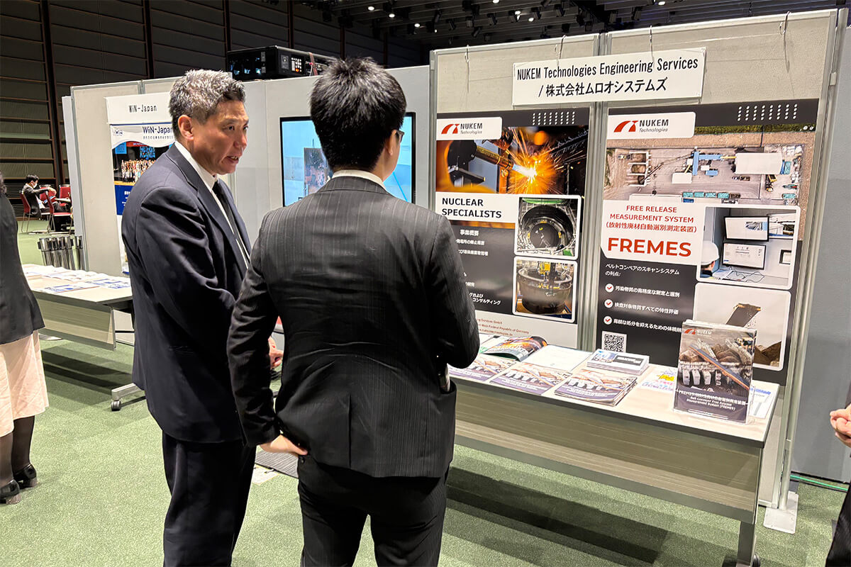 Zwei M&auml;nner im Anzug stehen an einem Messestand von NUKEM Technologies Engineering Services und sprechen &uuml;ber ausgestellte Informationen zu nuklearer Technik.