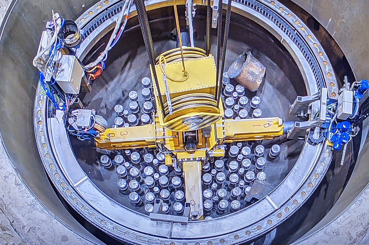 Luftaufnahme eines kreisf&ouml;rmigen, technischen Installationsbereichs mit einem gro&szlig;en, gelben Hebe- oder Montageger&auml;t in der Mitte, umgeben von zahlreichen zylindrischen Metallbeh&auml;ltern und elektrischen Kabeln.