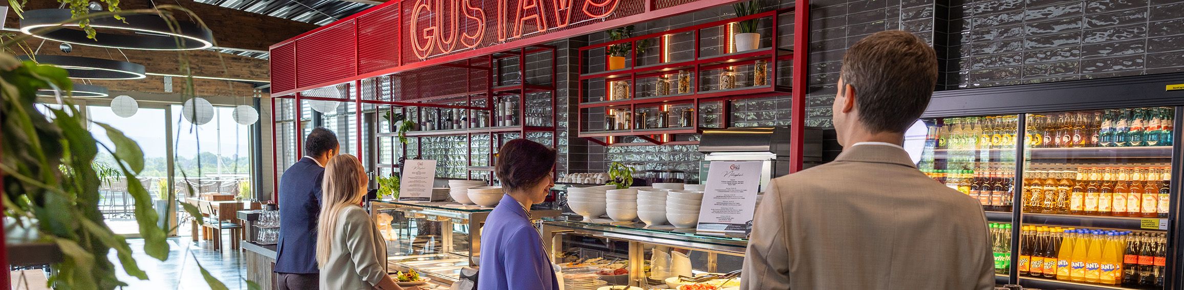 Mehrere Personen stehen in einer modernen Cafeteria an einem Selbstbedienungstresen mit Speisen und Getränken.