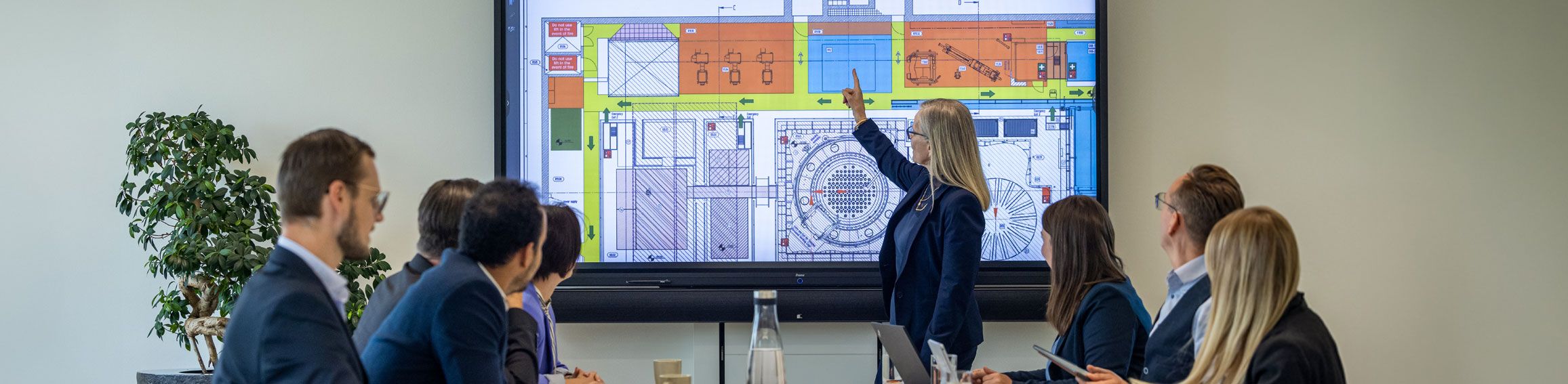 Eine Frau präsentiert einem Team einen farbigen technischen Plan auf einem großen Bildschirm in einem Konferenzraum.